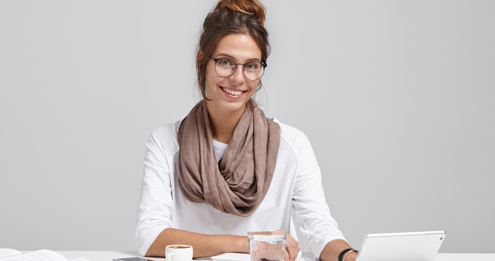 Cute female blogger types new interesting post at tablet, uses high speed internet connection, has confident and happy look, sits at work place alone. Elegant female lady makes financial report